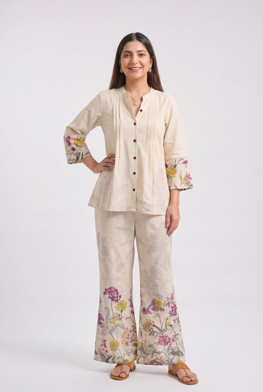 A woman modeling a two-piece cream-colored linen pajama set with floral prints on the cuffs and pant hems, photographed against a solid light gray background.