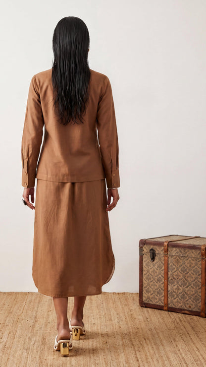 Woman in a brown suit standing next to a patterned suitcase on a wooden floor.