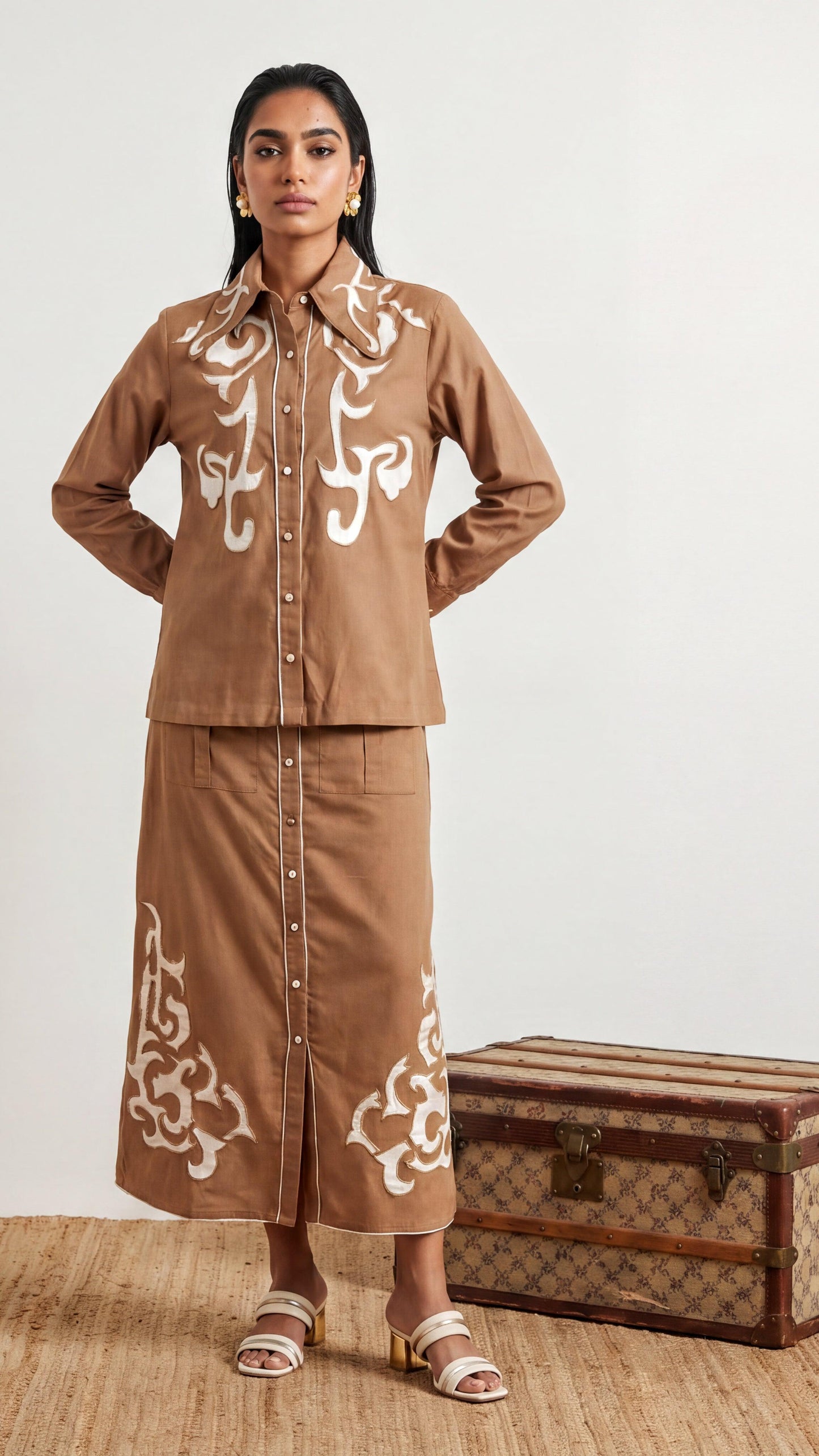 Woman wearing a brown outfit with white patterns, standing next to a vintage suitcase on a wooden floor.