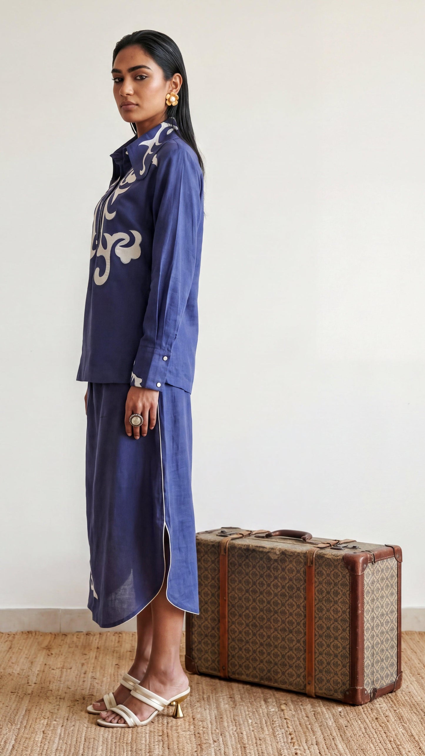 Woman in a blue outfit standing next to a vintage suitcase against a white wall.