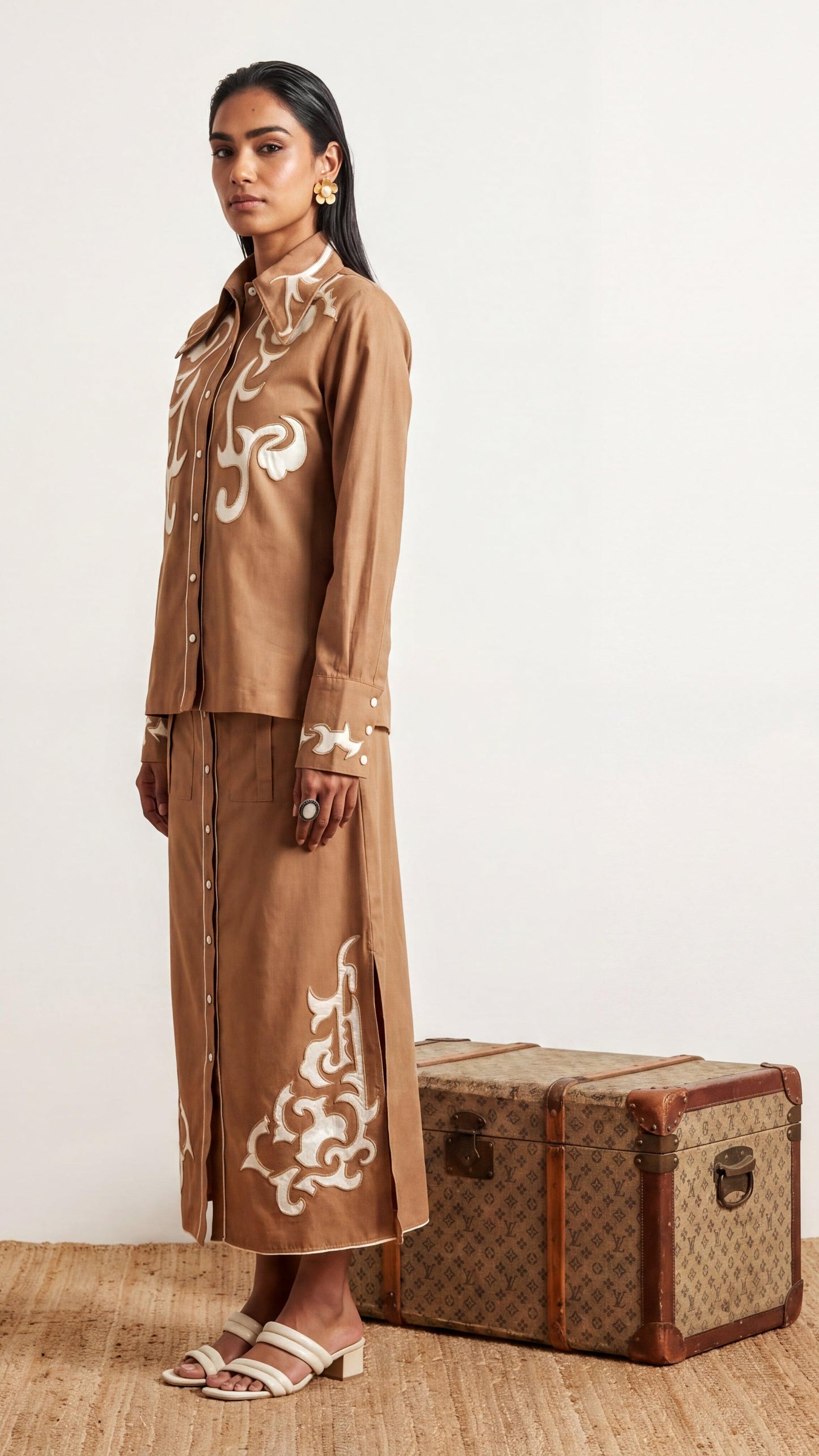 Woman in a brown outfit with white patterns standing next to a vintage suitcase on a beige background