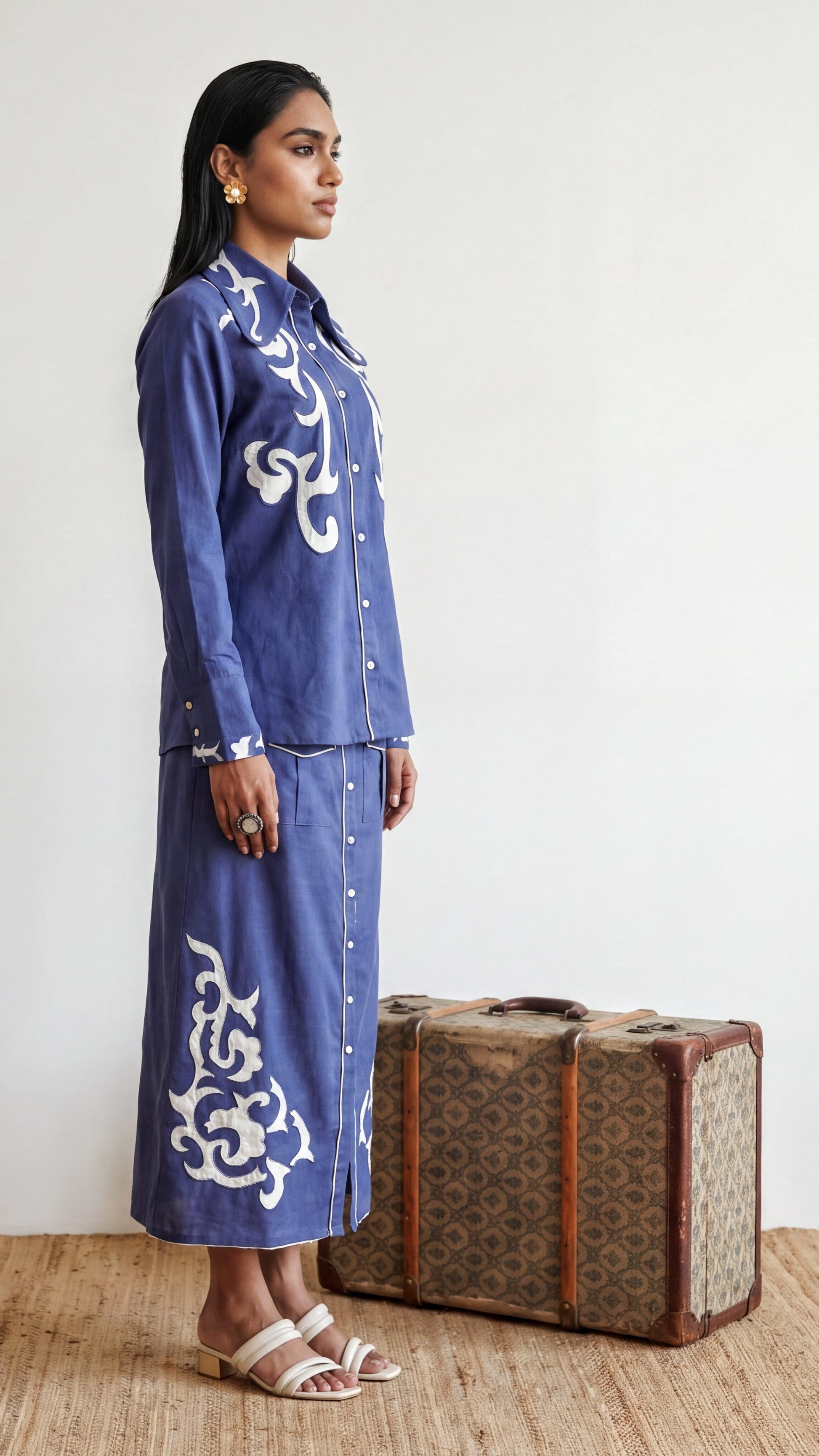 Woman in a blue embroidered outfit standing next to a vintage suitcase.