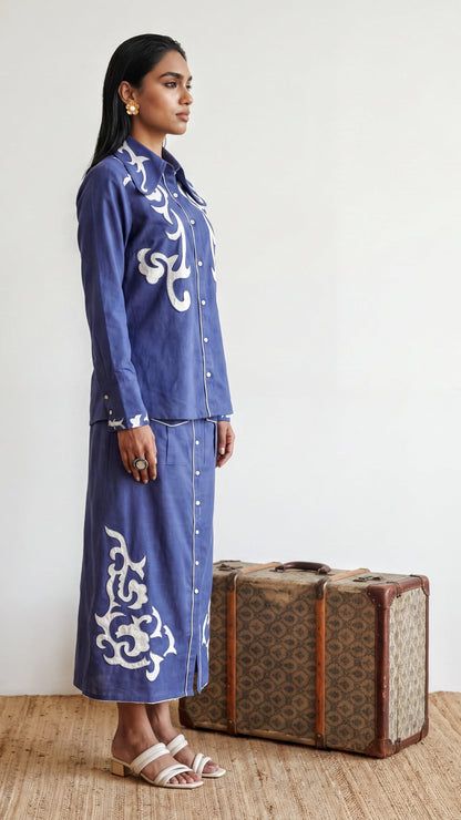 Woman in a blue embroidered outfit standing next to a vintage suitcase.