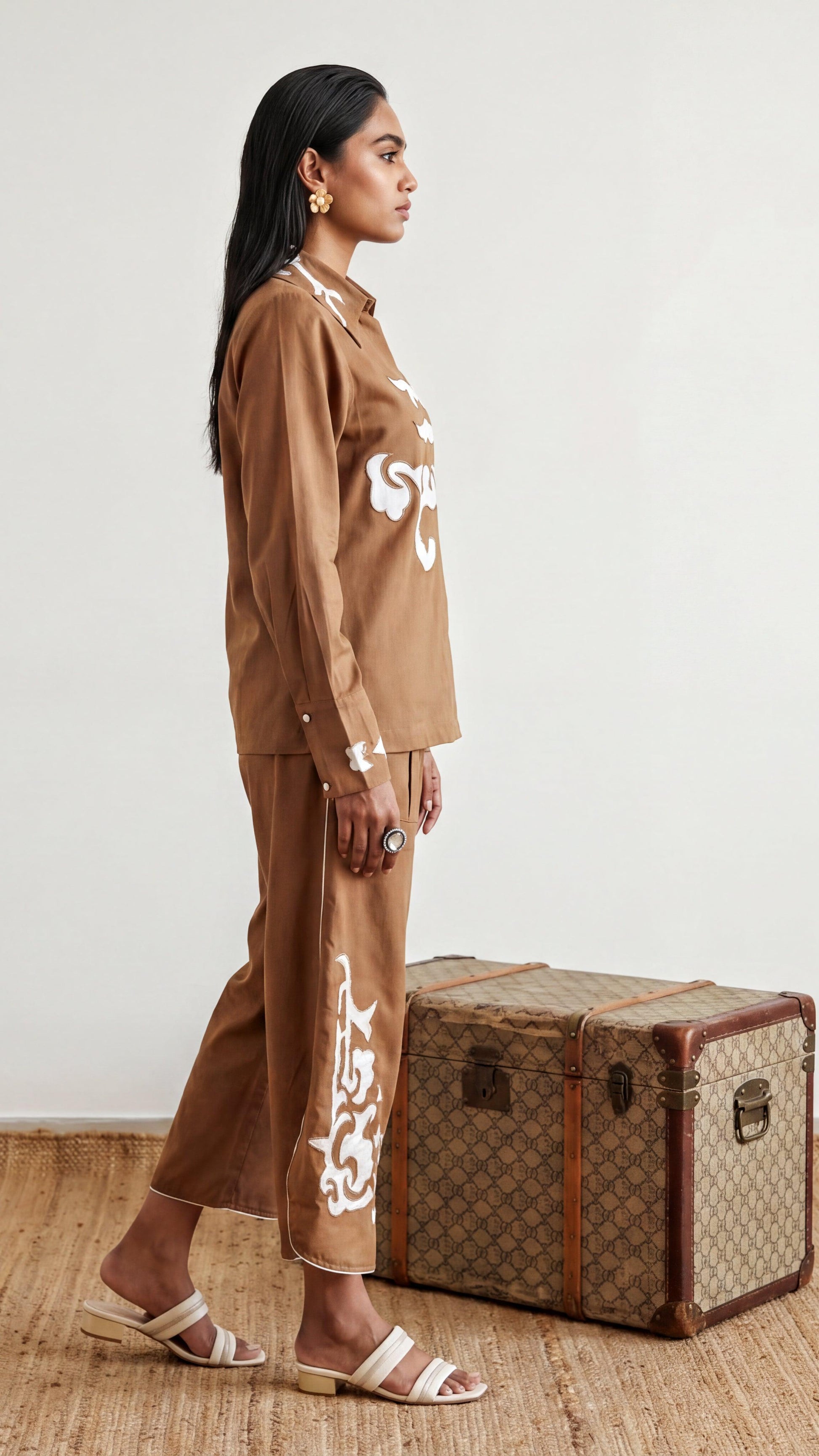 Woman in a brown outfit standing next to a vintage suitcase on a wooden floor.