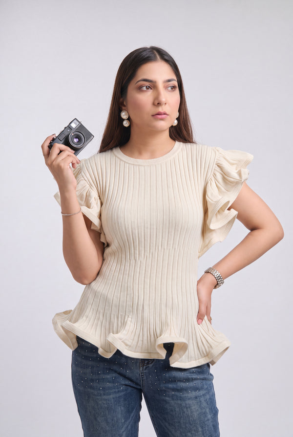 A woman modeling a cream-colored ribbed knit top with ruffled sleeves and a ruffled hem, paired with blue jeans, holding a small camera against a light gray background.