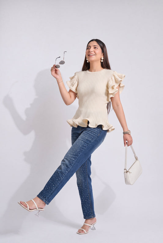 A woman posing in a cream ribbed knit top with ruffled sleeves and hem, paired with blue jeans and white sandals, holding a small white handbag and sunglasses against a light gray background.