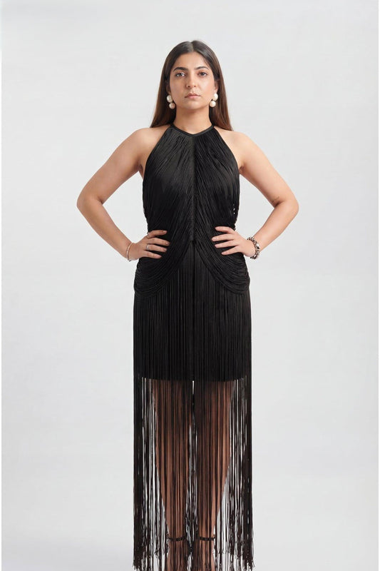 A woman modeling a sleeveless black halter dress with a pleated bodice and a long fringe skirt, photographed against a light gray studio background.