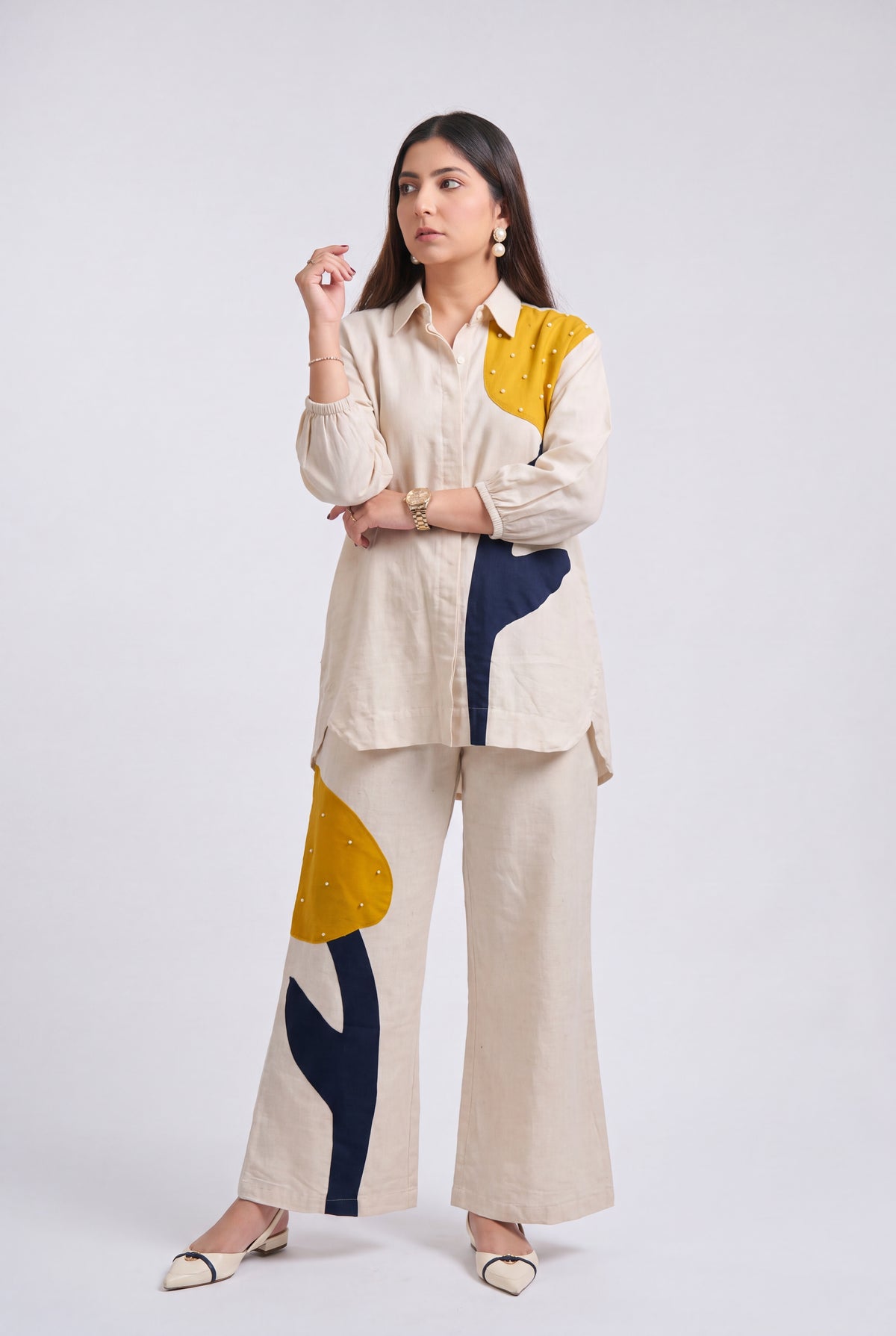 A woman modeling a matching two-piece outfit consisting of a cream-colored long-sleeve button-up shirt and wide-leg trousers, both decorated with abstract color-blocked patterns in mustard yellow, navy blue, and beige.