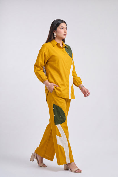 A woman modeling a mustard yellow two-piece outfit with green and white geometric patterned panels, standing against a light gray studio background.