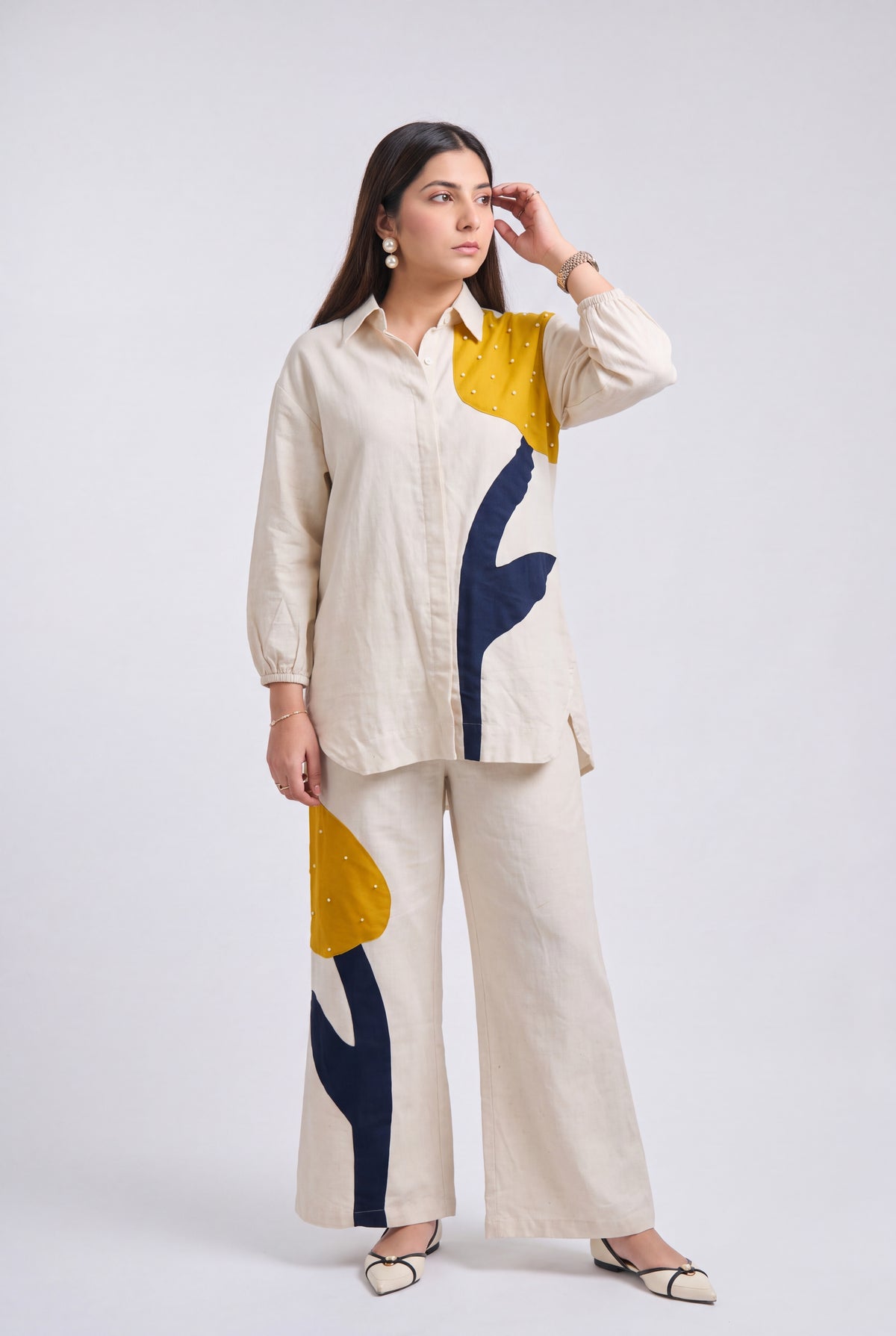 A woman modeling a matching two-piece linen set featuring a long-sleeve button-up shirt and wide-leg trousers, both in cream with abstract yellow, navy blue, and white color-blocked patterns.