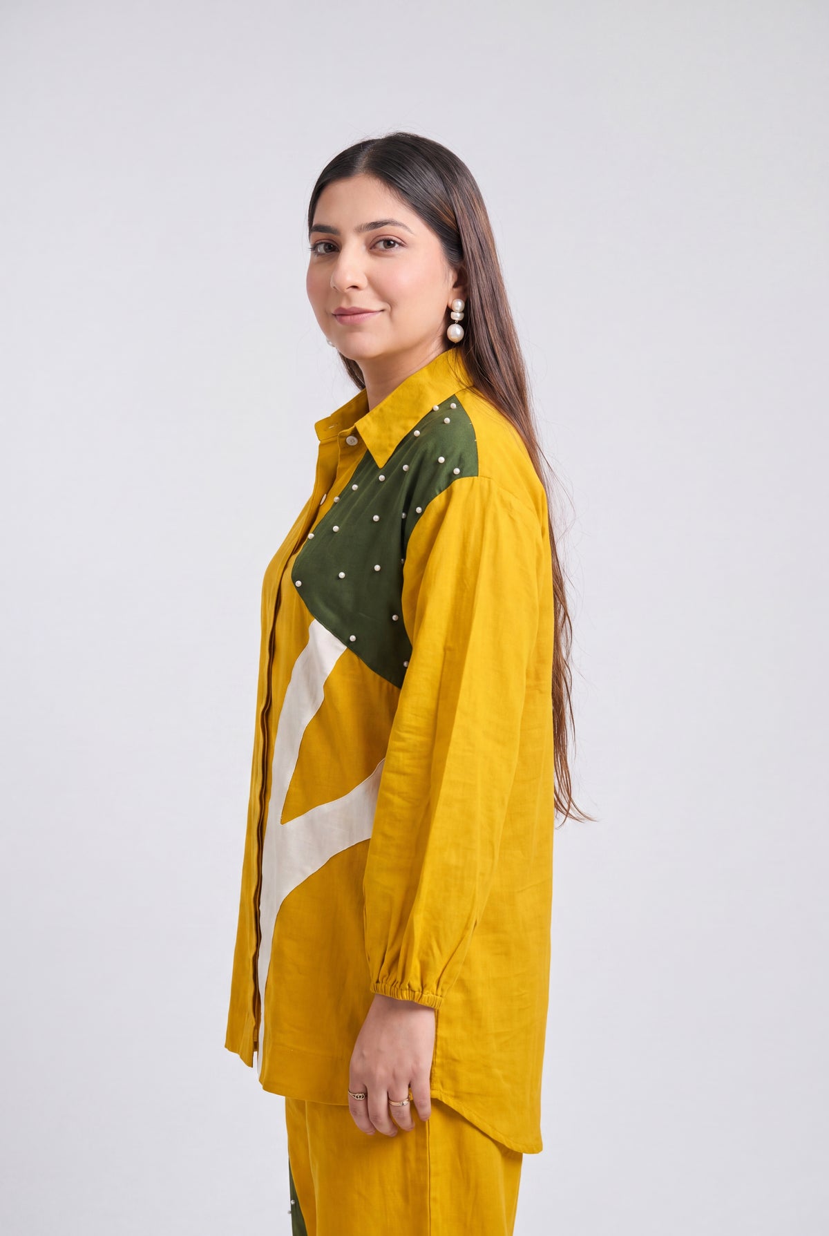 A woman modeling a mustard yellow long-sleeve shirt with green and white patterned panels on the chest, paired with matching yellow pants, against a light gray studio background.