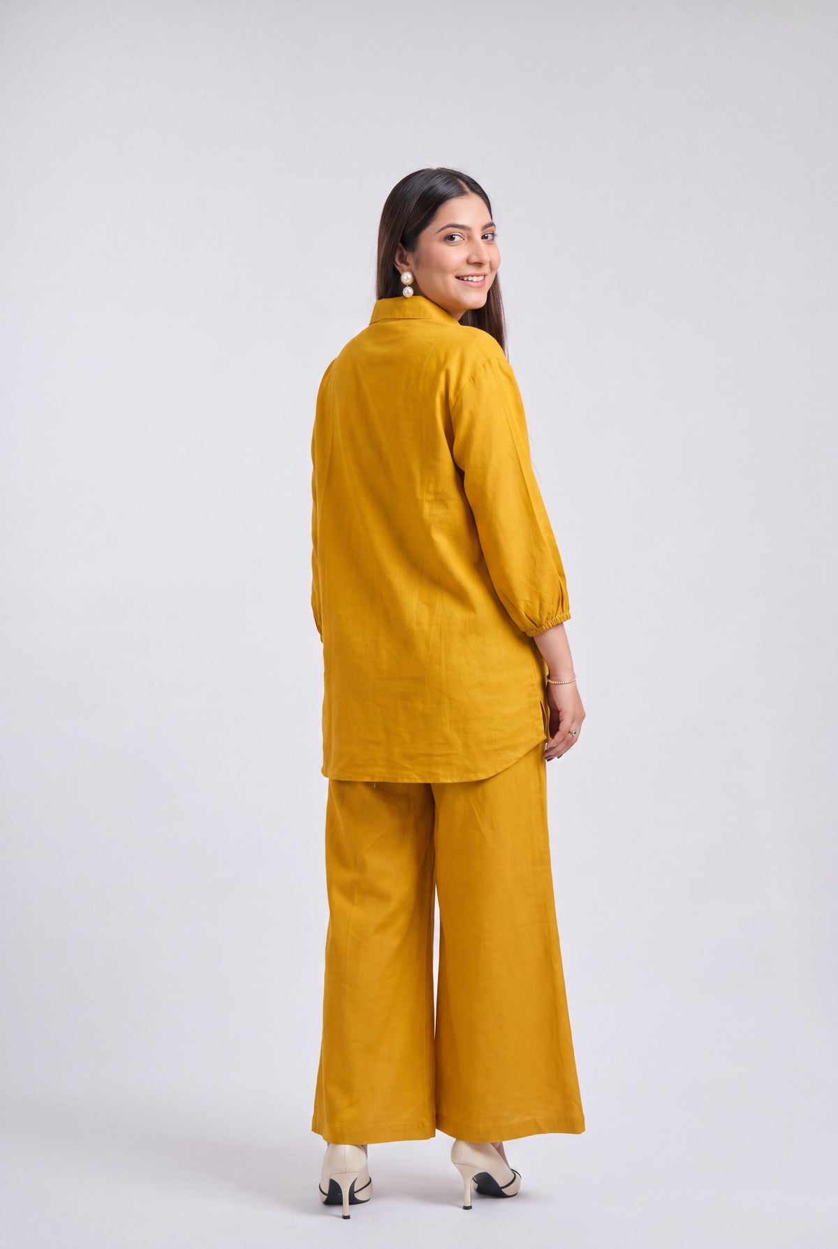 A woman modeling a mustard yellow two-piece linen set, shown from the back as she looks over her shoulder. The set includes a short-sleeved tunic and matching wide-leg trousers, photographed against a neutral light gray studio background.