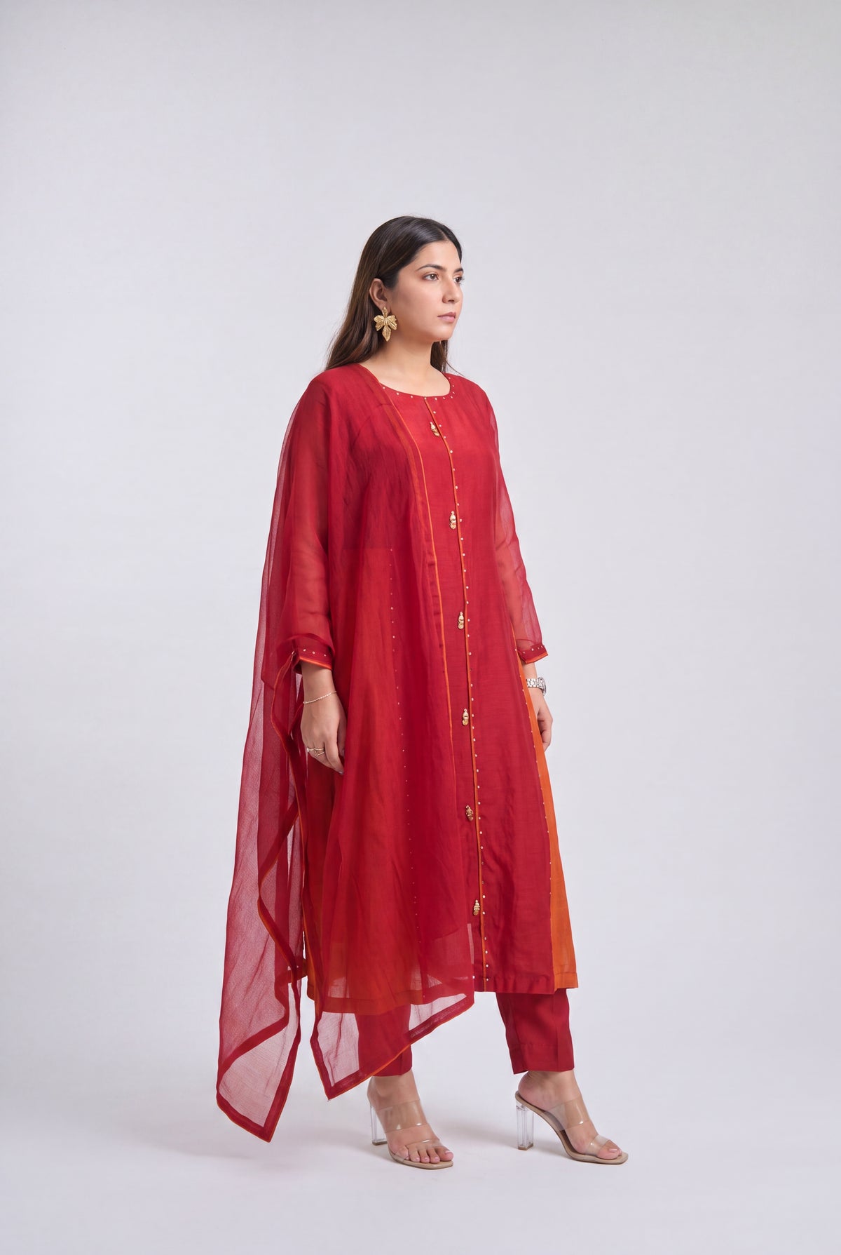 A woman modeling a red three-piece traditional outfit consisting of a long kurta with gold button details, matching pants, and a sheer red dupatta, set against a plain light gray studio background.