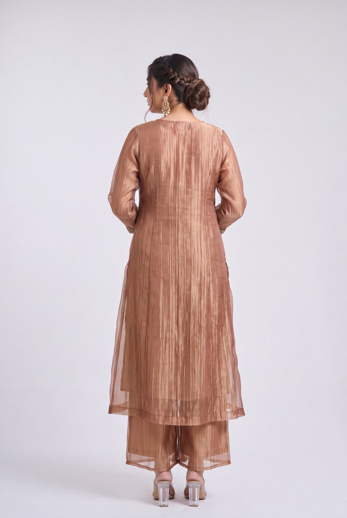 A woman viewed from the back wearing a metallic bronze traditional outfit with a long kurta and matching pants, standing against a light gray studio background.