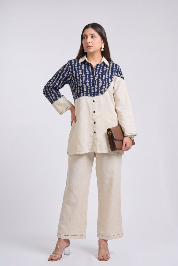 A woman models a two-tone shirt and trouser set against a plain light gray studio background. The shirt has a navy blue and cream patterned upper section and solid cream lower section, paired with matching cream wide-leg trousers.