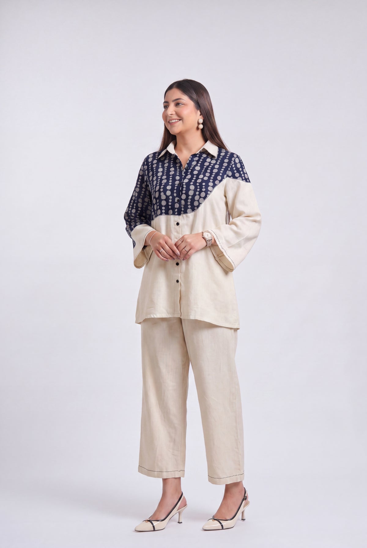 A woman modeling a color-blocked shirt and trouser set against a light gray studio background. The shirt features a navy blue and white patterned yoke with solid cream sleeves and collar, paired with matching cream wide-leg cropped trousers.