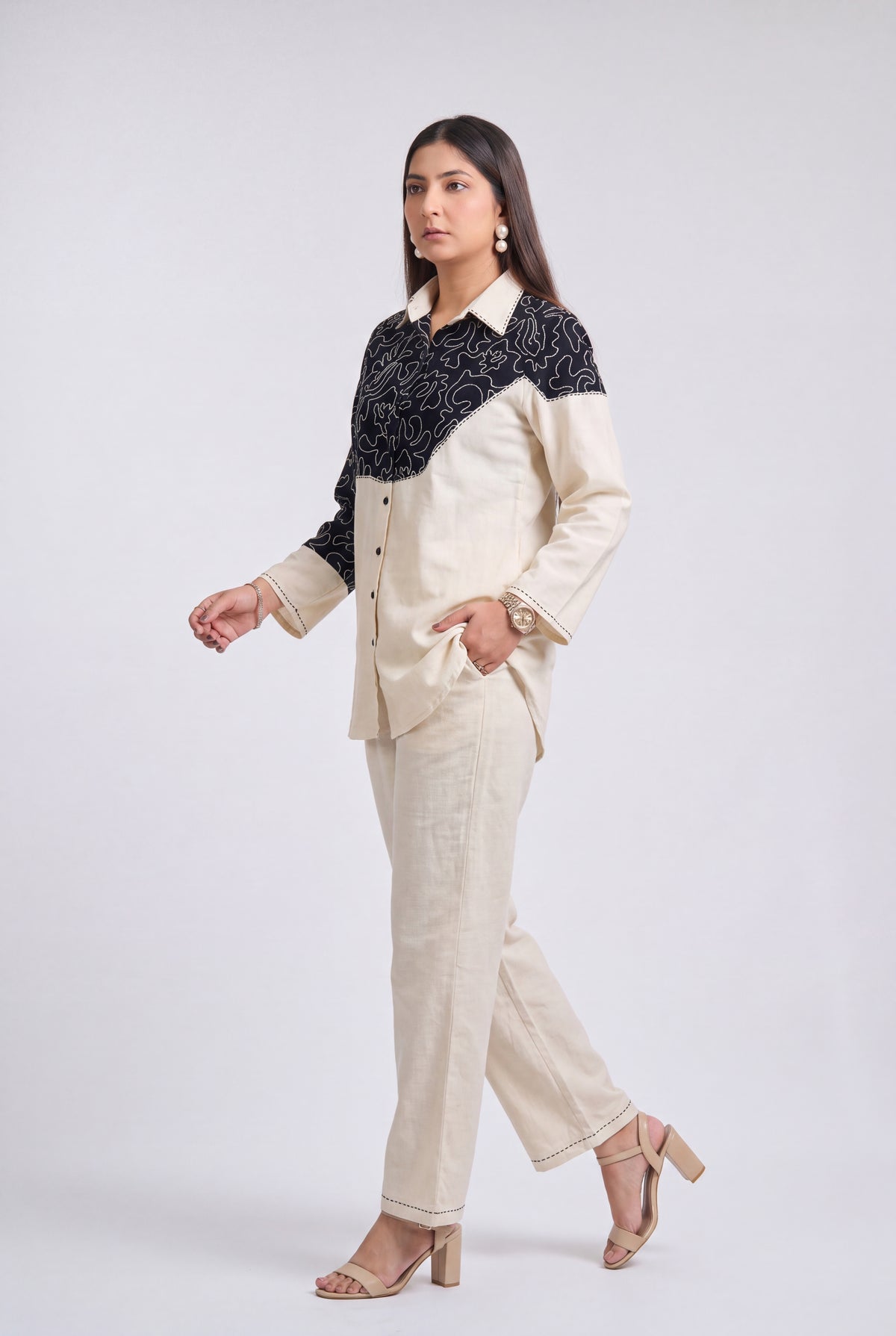 A woman modeling a two-piece suit featuring a cream-colored linen pantsuit with a contrasting black embroidered yoke on the shirt jacket, posed against a light gray studio background.