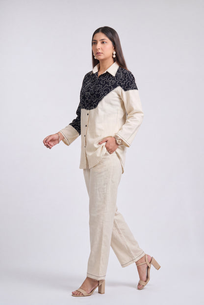 A woman modeling a two-piece suit featuring a cream-colored linen pantsuit with a contrasting black embroidered yoke on the shirt jacket, posed against a light gray studio background.