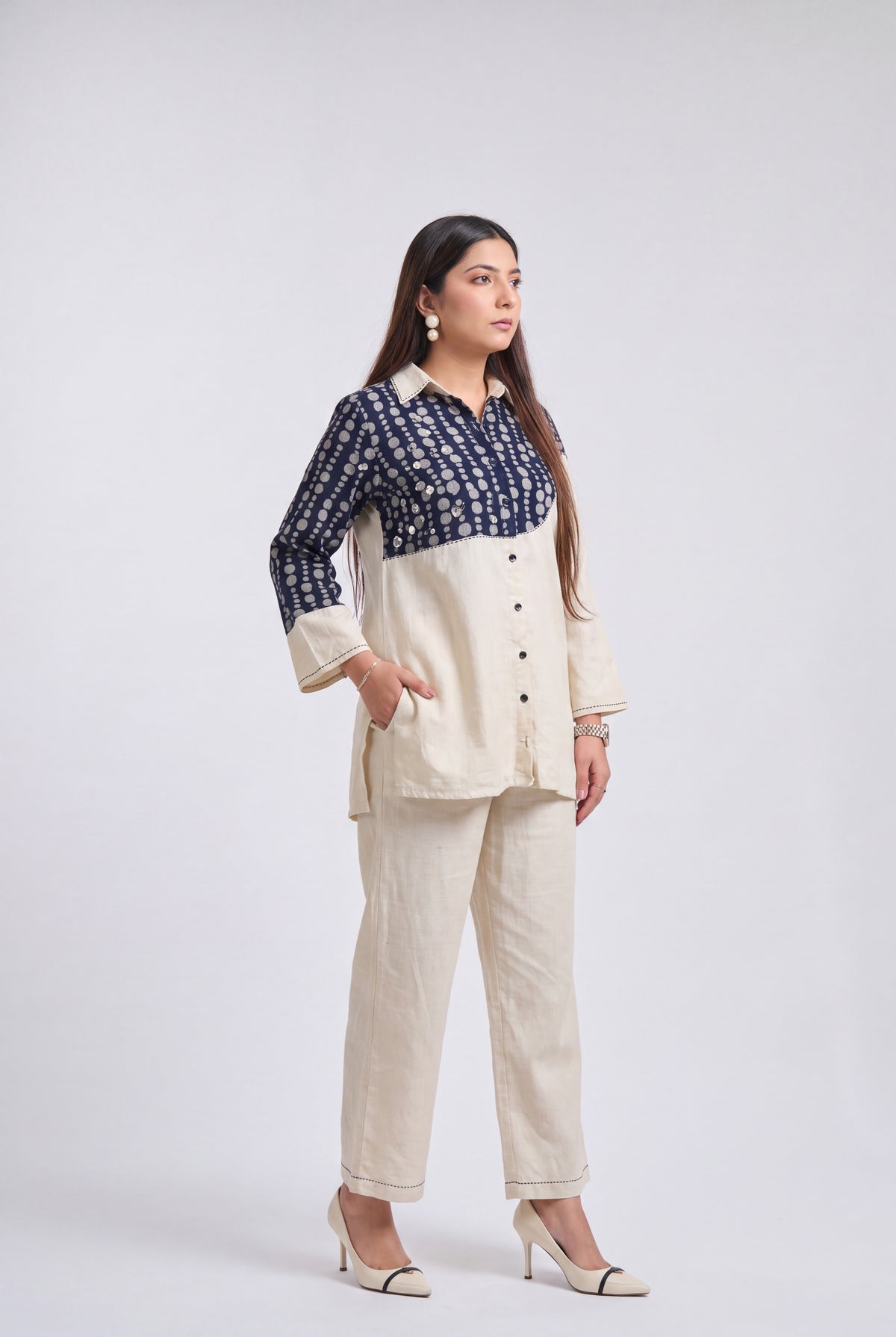 A woman modeling a two-piece outfit consisting of a cream-colored linen pants set and a matching cream shirt with a navy blue patterned yoke, styled with beige heels against a light gray studio background.