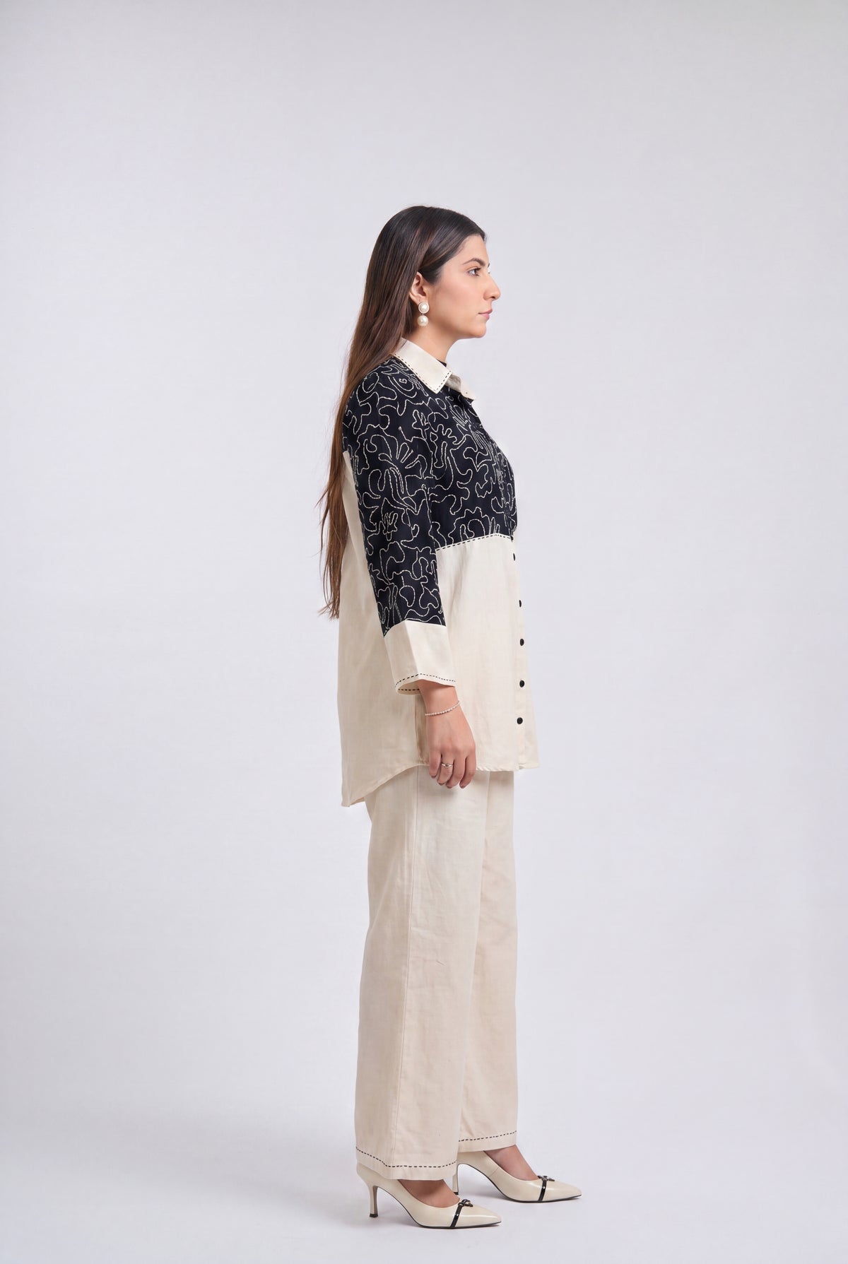 A woman stands in profile wearing a two-tone outfit consisting of a cream and black patterned button-front top with wide-leg cream pants, photographed against a plain light gray studio background.