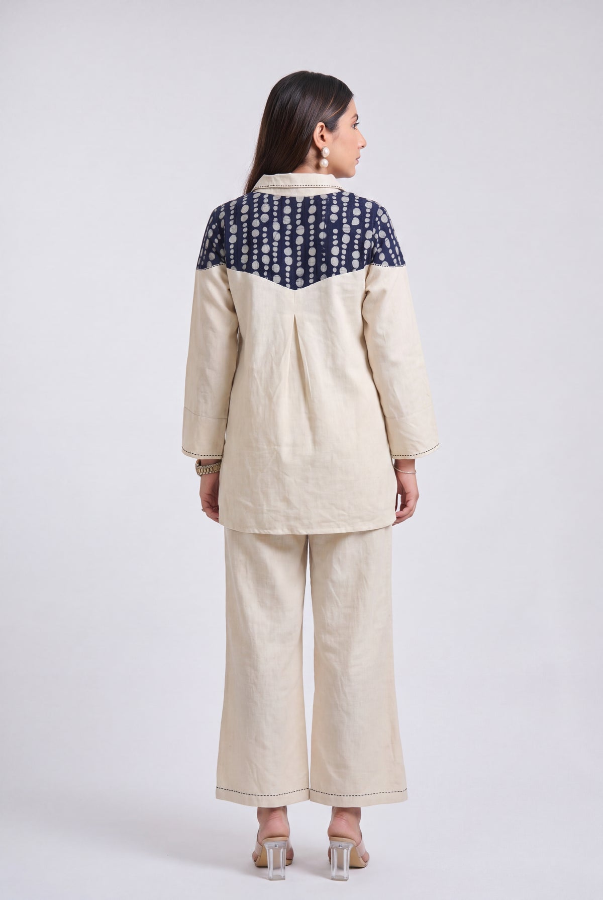 A woman shown from the back wearing a two-piece linen outfit. The set consists of a cream-colored long-sleeve top with a navy blue and white patterned yoke, paired with matching cream wide-leg trousers, against a light gray studio background.