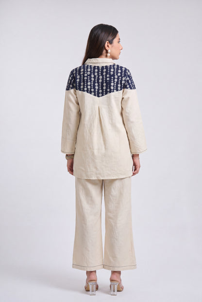 A woman shown from the back wearing a two-piece linen outfit. The set consists of a cream-colored long-sleeve top with a navy blue and white patterned yoke, paired with matching cream wide-leg trousers, against a light gray studio background.