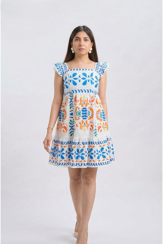 A woman modeling a short white dress with colorful blue and orange floral embroidery, featuring ruffled cap sleeves and a tiered skirt, set against a plain light gray background.