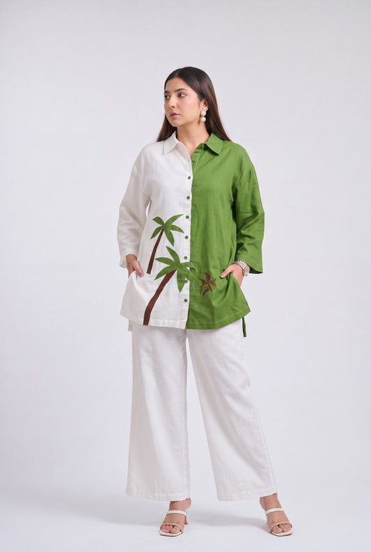 A woman modeling a two-tone button-up shirt with white on one side and olive green on the other, featuring a palm tree embroidery on the white side, paired with white wide-leg pants against a light gray studio background.