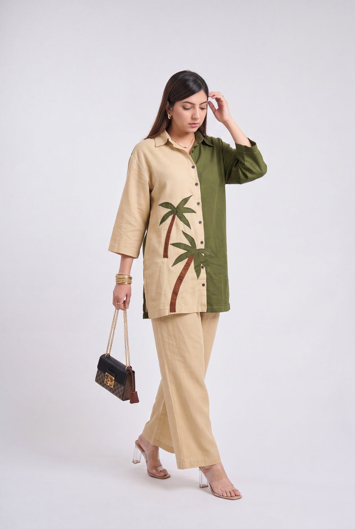 A woman models a two-tone beige and olive green kurta set with embroidered palm trees, paired with matching beige trousers and holding a small black handbag against a light gray studio background.