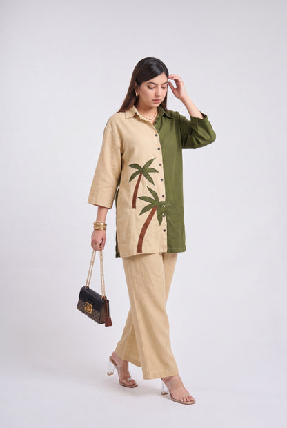 A woman models a two-tone beige and olive green kurta set with embroidered palm trees, paired with matching beige trousers and holding a small black handbag against a light gray studio background.