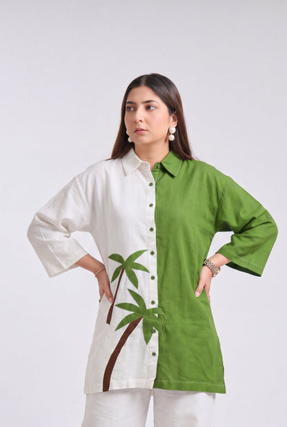 A woman modeling a split-color button-up shirt with one half in white and the other in green, featuring a palm tree graphic on the white side, paired with white wide-leg pants and beige flat shoes, against a light gray studio background.
