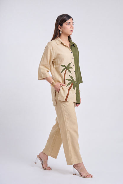 A woman models a two-piece linen set consisting of a beige short-sleeve button-up shirt with green palm tree embroidery and matching beige wide-leg trousers, styled with an olive green cardigan and clear high-heeled sandals against a light gray studio background.