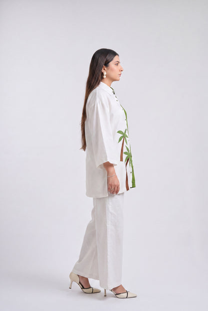 A woman stands in profile wearing a white two-piece outfit with a green and brown palm tree print on the jacket, paired with white wide-leg pants and beige high heels, against a light gray studio background.