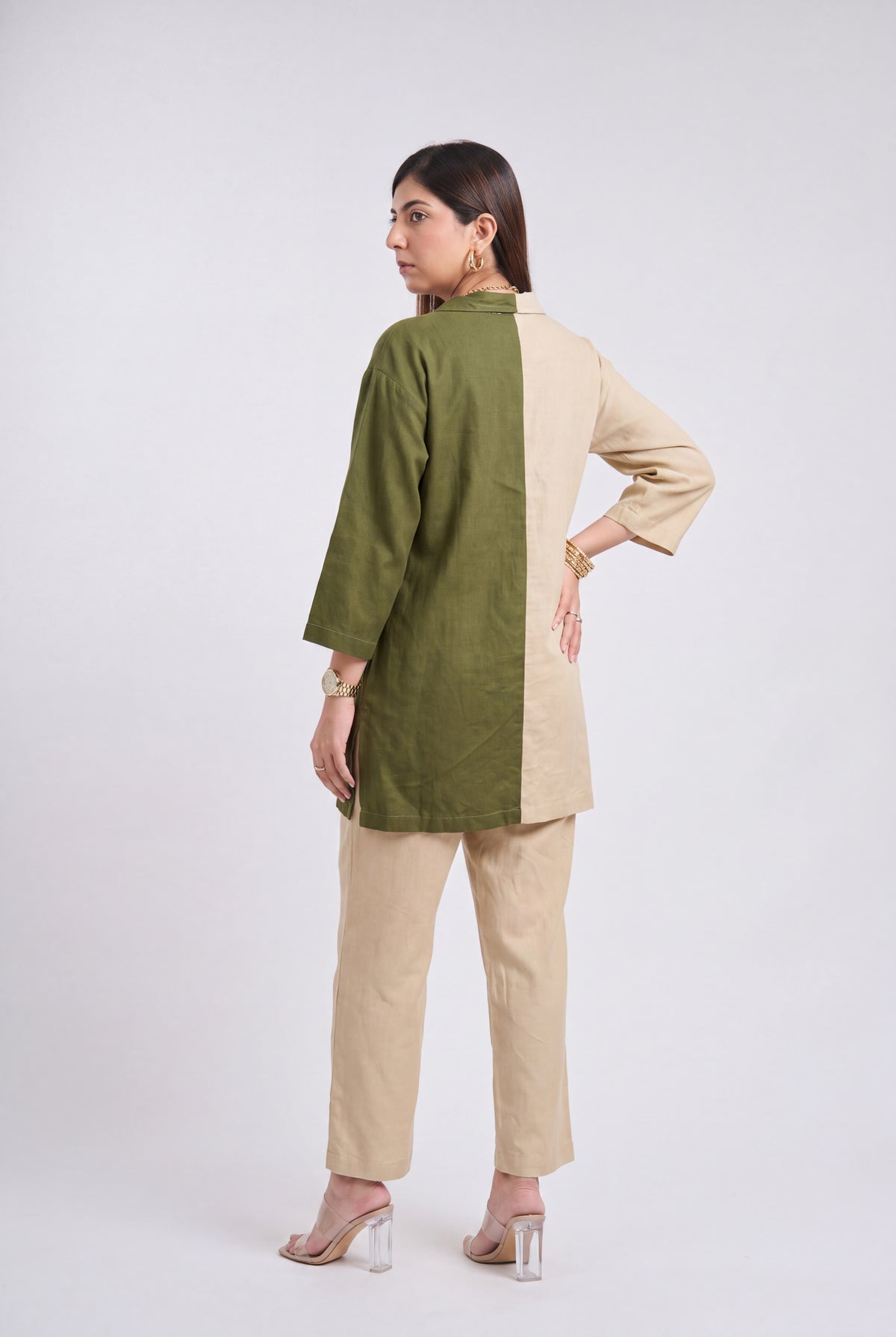 A woman modeling a two-tone olive green and beige tunic top with matching beige trousers, shown from a rear three-quarter view against a light gray studio background.