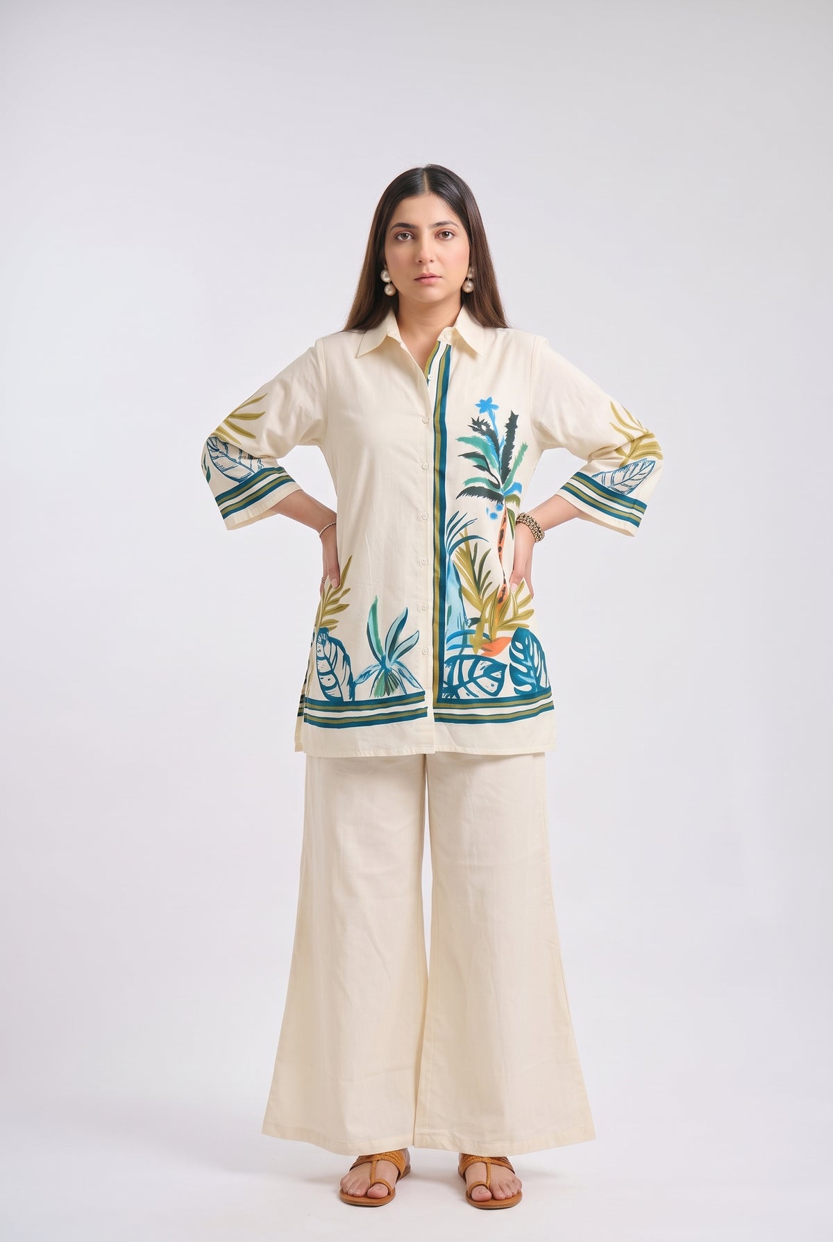 A woman modeling a cream-colored two-piece outfit consisting of a long-sleeve button-up shirt with colorful tropical embroidery and matching wide-leg trousers, set against a plain light gray studio background.