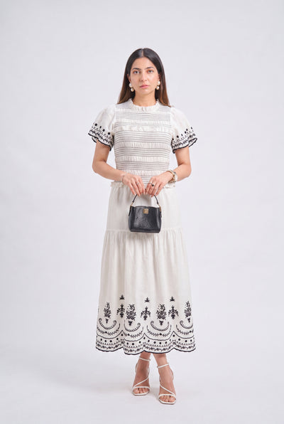 A woman modeling a cream-colored tiered midi dress with black embroidered details on the bodice, sleeves, and hem, holding a small black handbag against a light gray studio background.