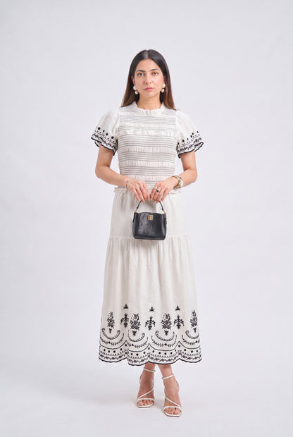 A woman modeling a cream-colored tiered midi dress with black embroidered details on the bodice, sleeves, and hem, holding a small black handbag against a light gray studio background.