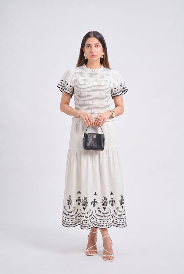 A woman modeling a cream-colored tiered midi dress with black embroidered details on the bodice, sleeves, and hem, holding a small black handbag against a light gray studio background.