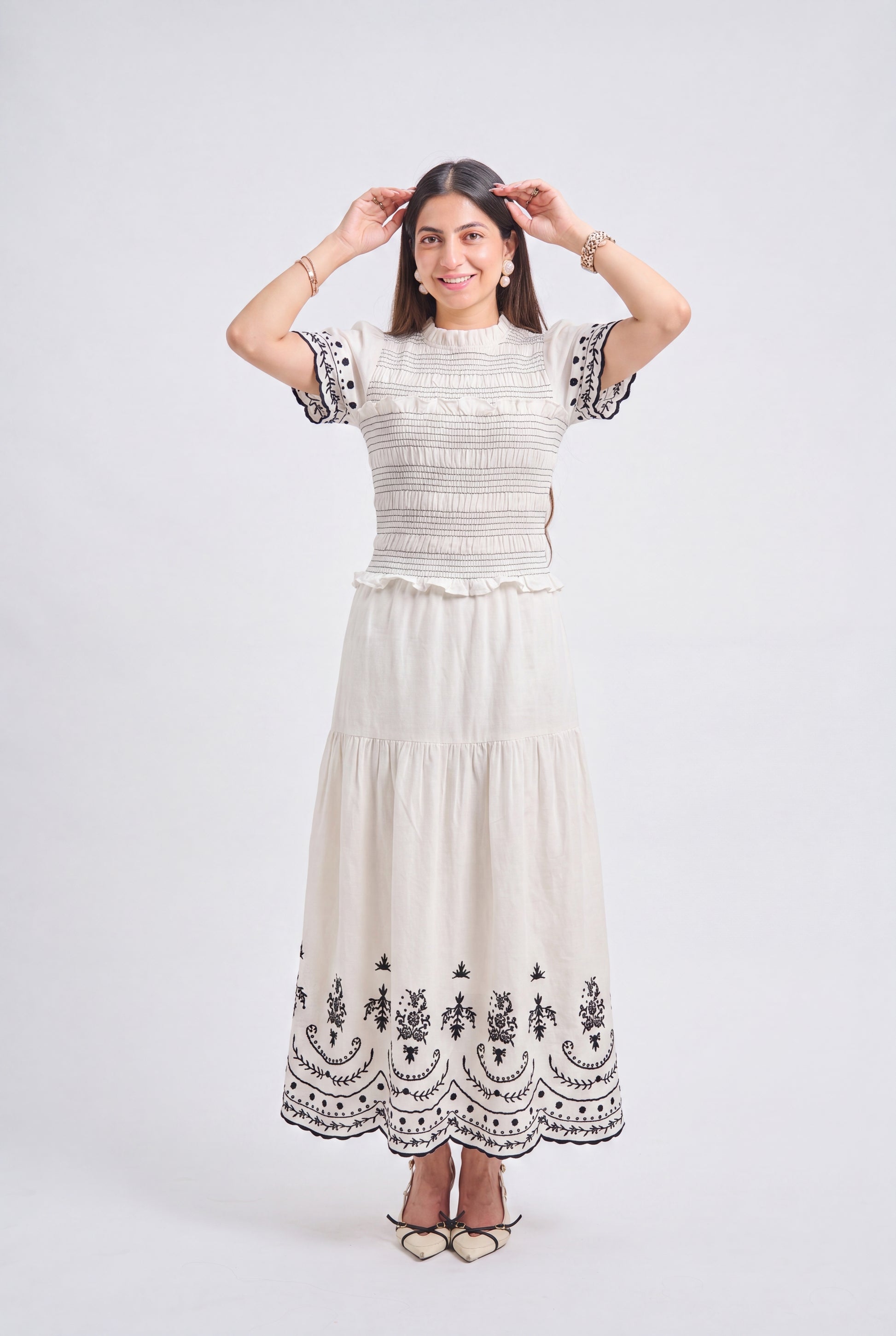 A woman modeling a cream-colored two-piece dress set featuring a smocked short-sleeve top and a tiered maxi skirt with black floral embroidery, set against a plain light gray studio background.