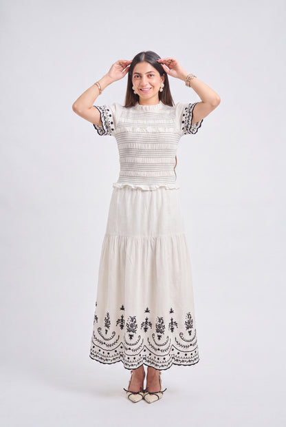 A woman modeling a cream-colored two-piece dress set featuring a smocked short-sleeve top and a tiered maxi skirt with black floral embroidery, set against a plain light gray studio background.