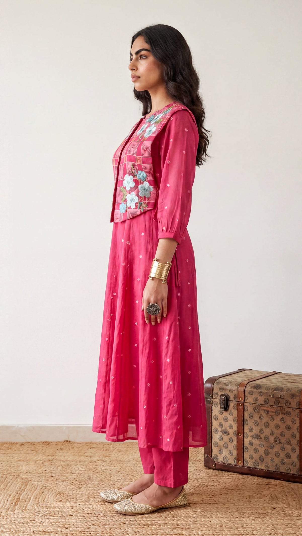 Woman in a pink traditional outfit standing next to a vintage suitcase on a wooden floor.