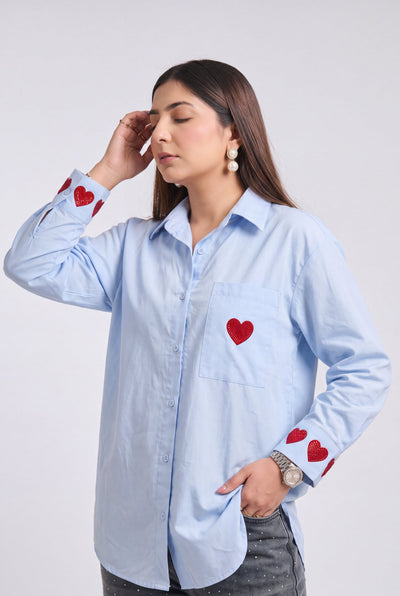 A woman modeling a light blue long-sleeve button-up shirt with red heart embroidery details on the chest pocket and cuffs, paired with distressed gray jeans, against a plain light gray background.
