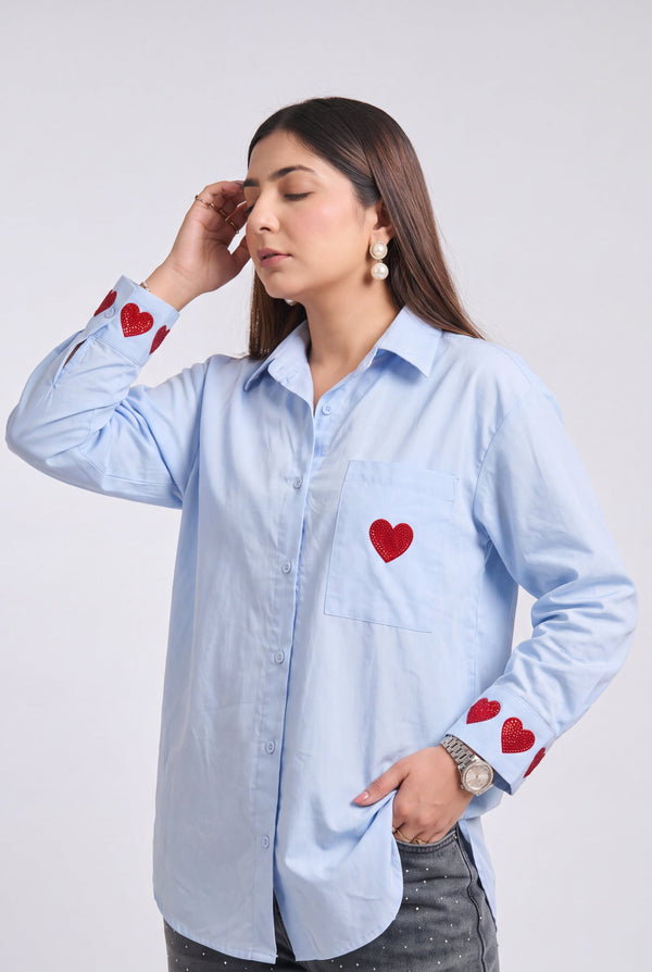 A woman modeling a light blue long-sleeve button-up shirt with red heart embroidery details on the chest pocket and cuffs, paired with distressed gray jeans, against a plain light gray background.