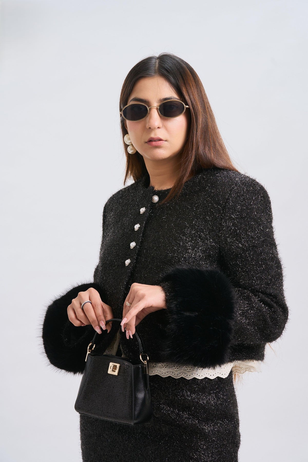 Woman wearing a black textured jacket with fur trim, holding a black handbag against a white background.