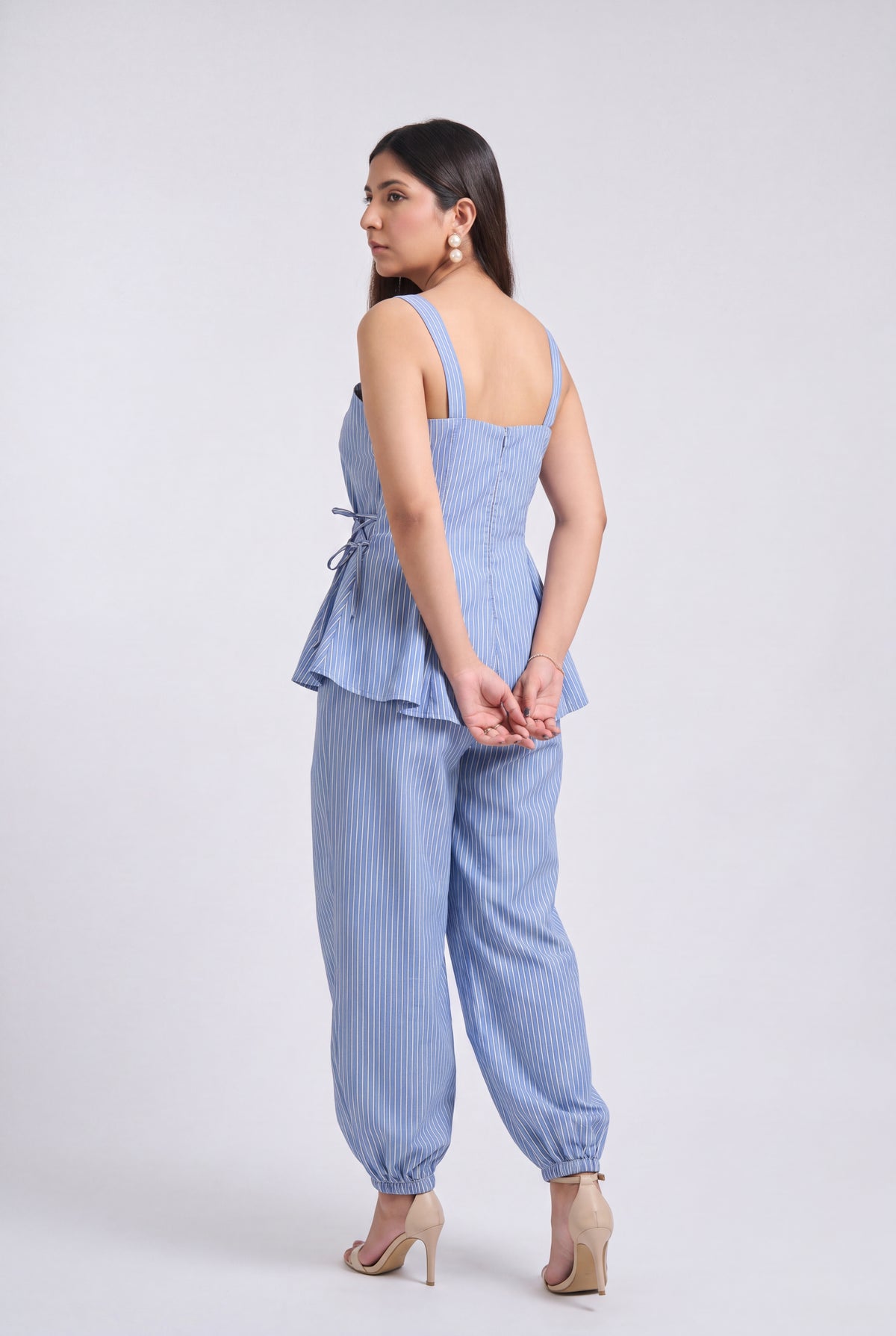 A woman modeling a light blue and white vertically striped two-piece set consisting of a sleeveless peplum top and matching wide-leg pants, shown from a rear three-quarter angle against a plain light gray studio background.