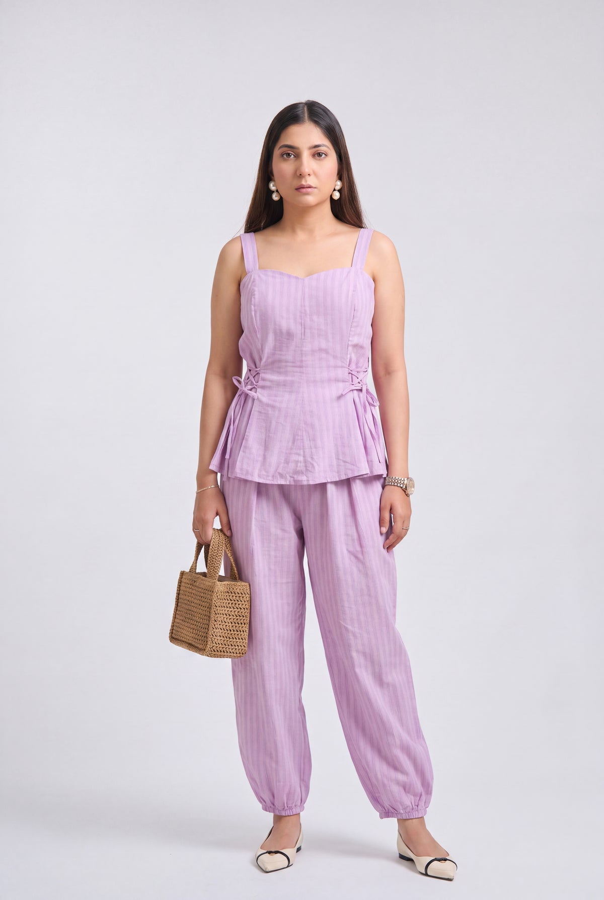 A woman modeling a light purple/lavender two-piece set consisting of a sleeveless peplum top and matching wide-leg pants, holding a small woven tan handbag against a plain light gray studio background.