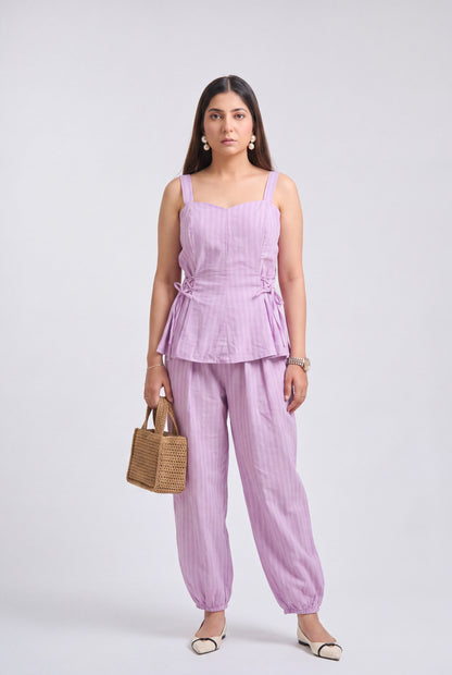 A woman modeling a light purple/lavender two-piece set consisting of a sleeveless peplum top and matching wide-leg pants, holding a small woven tan handbag against a plain light gray studio background.