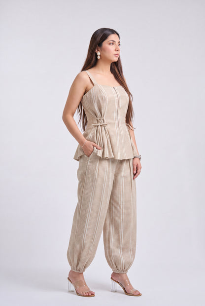 A woman modeling a light beige two-piece set consisting of a sleeveless peplum-style top with thin straps and a matching pair of loose, wide-leg pants, photographed against a plain light gray studio background.