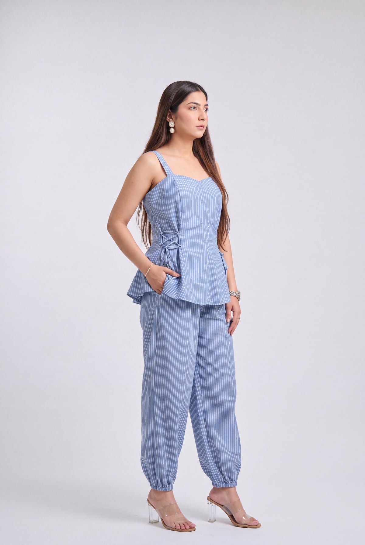 A woman modeling a light blue and white vertically striped two-piece set consisting of a sleeveless peplum top and matching wide-leg pants, photographed against a neutral light gray studio background.