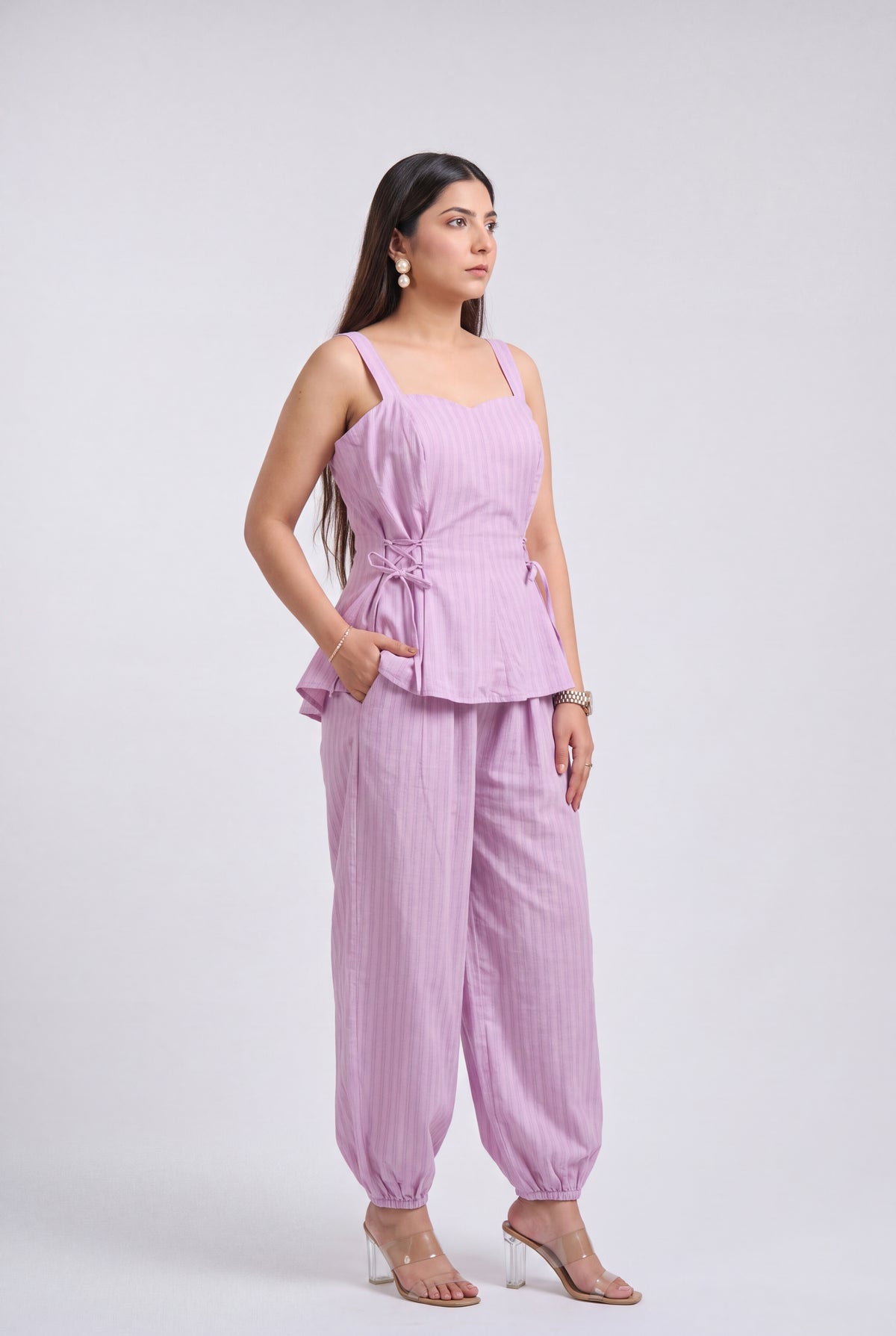 A woman modeling a light purple/lavender two-piece set consisting of a sleeveless peplum top with a side tie and matching wide-leg pants, photographed against a plain light gray studio background.